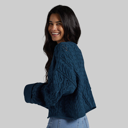 A woman with long dark hair is shown from the back and side, wearing a textured navy blue cable-knit cropped sweater with ribbed cuffs, paired with light blue jeans, against a solid light gray background.