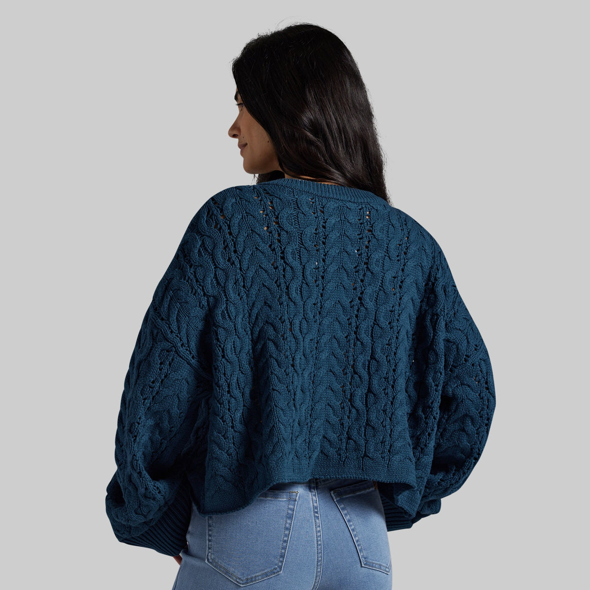 A woman shown from the back wearing a dark teal cable-knit cropped sweater and light blue jeans against a solid gray studio background.
