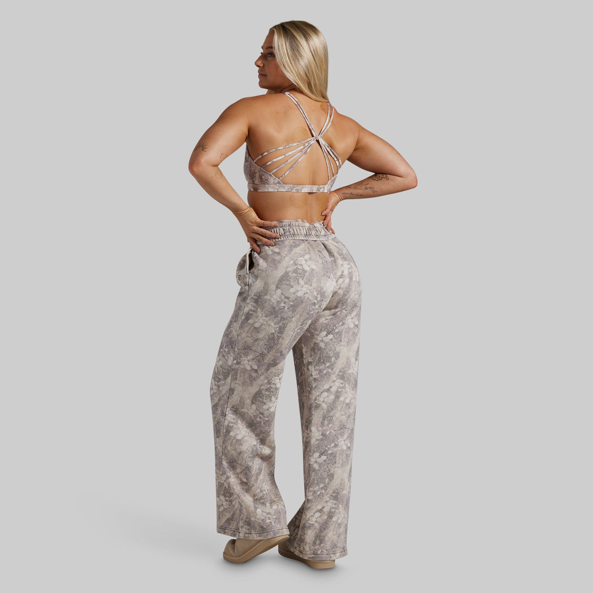 A woman is shown from the back wearing a matching two-piece lounge set consisting of a strappy sports bra and high-waisted, wide-leg pants in a gray and beige marble pattern, set against a solid gray background.