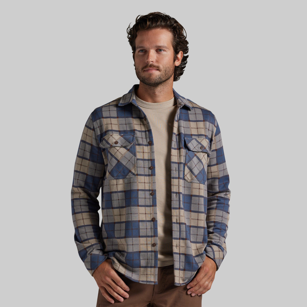 A man wearing a blue and beige plaid button-up flannel shirt over a beige t-shirt and brown pants, photographed against a solid gray studio background.