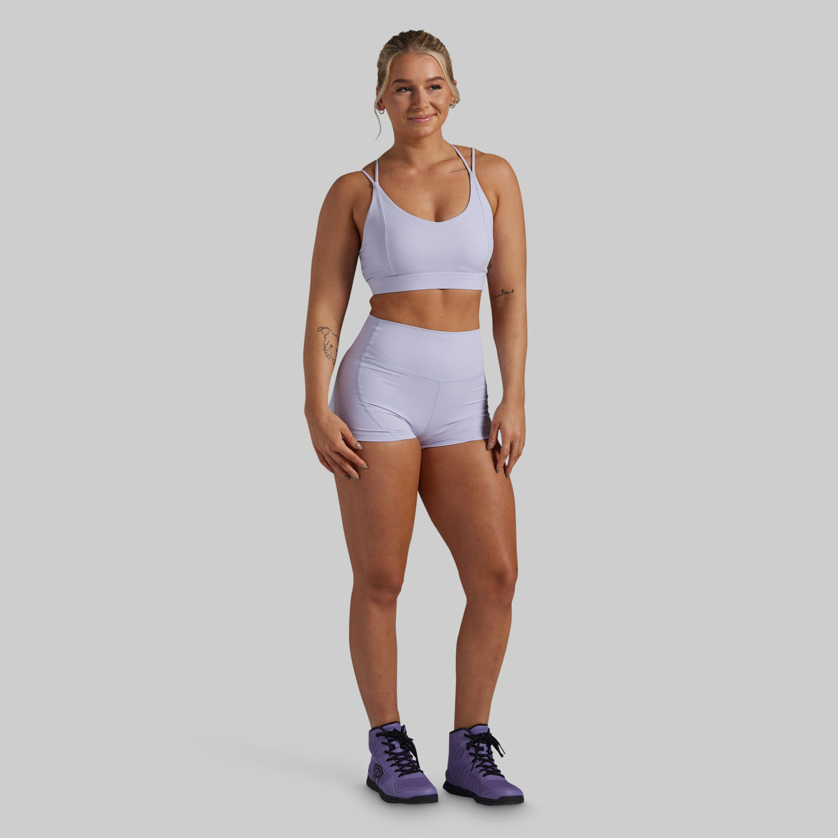 A woman modeling a light lavender sports bra and matching high-waisted athletic shorts against a solid gray studio background.