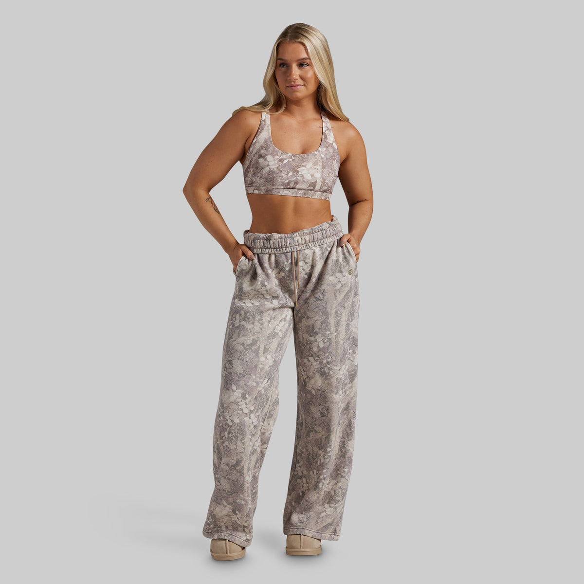 A woman modeling a matching two-piece activewear set featuring a beige and gray camouflage print sports bra and high-waisted, wide-leg sweatpants, set against a solid gray studio background.