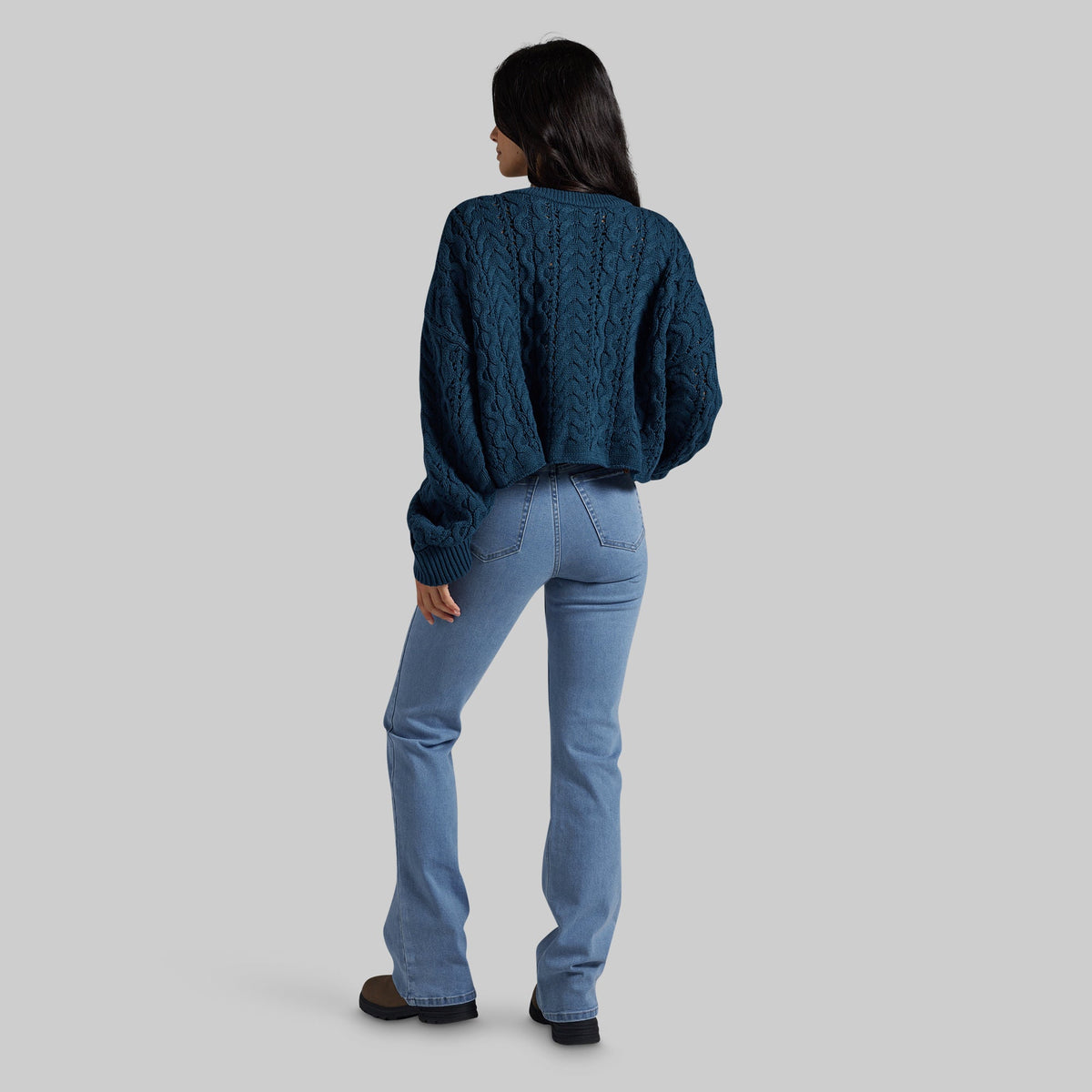 A woman shown from the back wearing a dark teal cable-knit cropped sweater and light blue jeans, standing against a solid gray studio background.