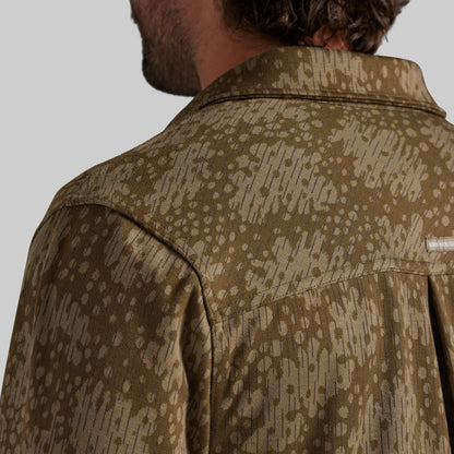 Close-up back view of a man wearing a collared shirt jacket with an abstract camouflage pattern in olive green and beige.