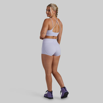 A woman modeling a light purple/lavender activewear set consisting of a sports bra and high-waisted shorts, shown from a rear three-quarter view against a solid gray studio background.