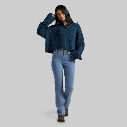 A woman modeling a dark teal blue cable-knit cropped henley sweater with light-wash blue jeans and brown boots, standing against a solid gray studio background.