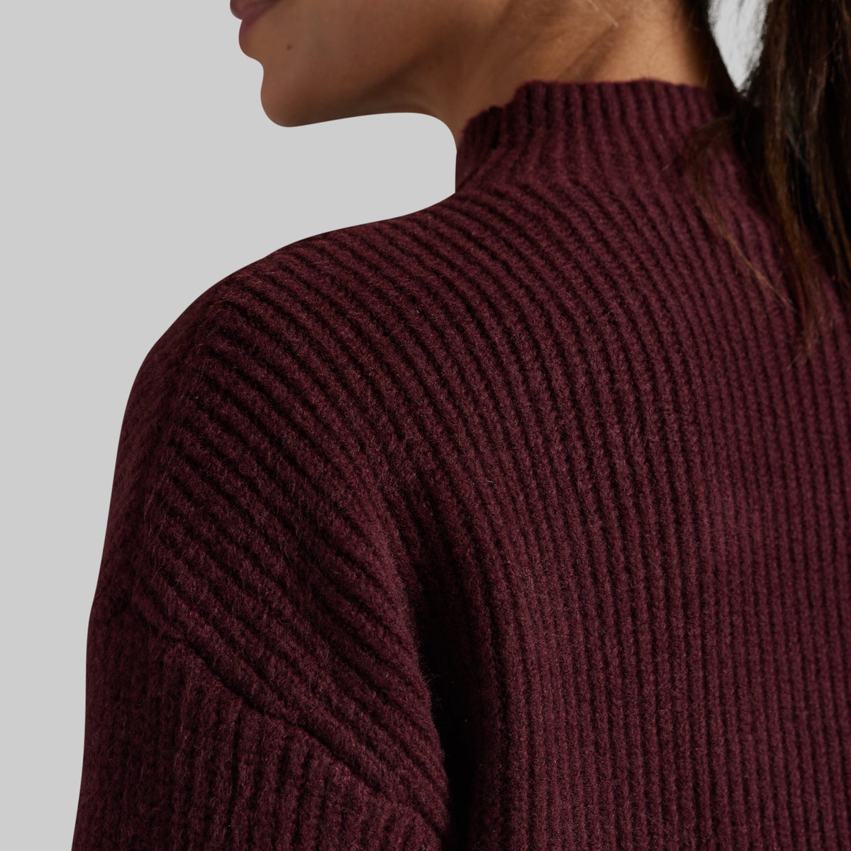Close-up of a person wearing a ribbed knit mock neck sweater in deep burgundy against a light gray background.