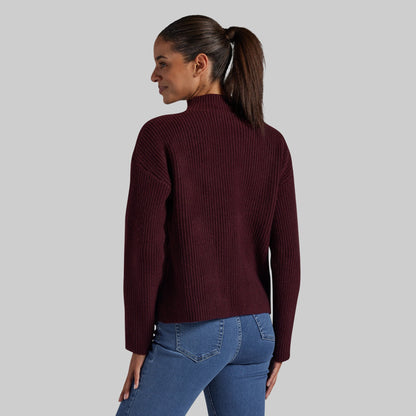 A woman shown from the back wearing a ribbed mock neck sweater in burgundy and blue jeans, set against a solid light gray background.