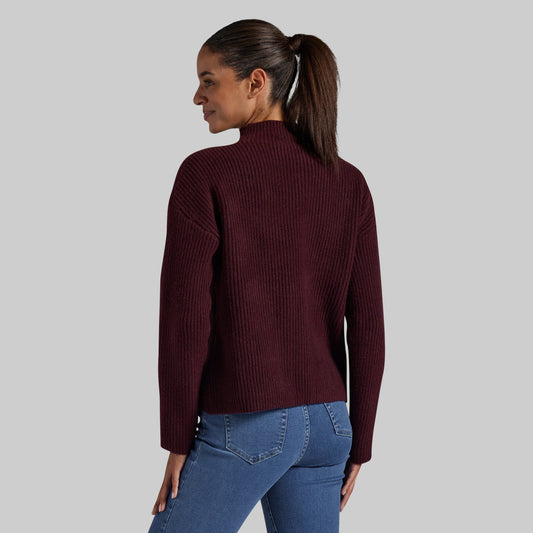A woman shown from the back wearing a ribbed mock neck sweater in burgundy and blue jeans, set against a solid light gray background.