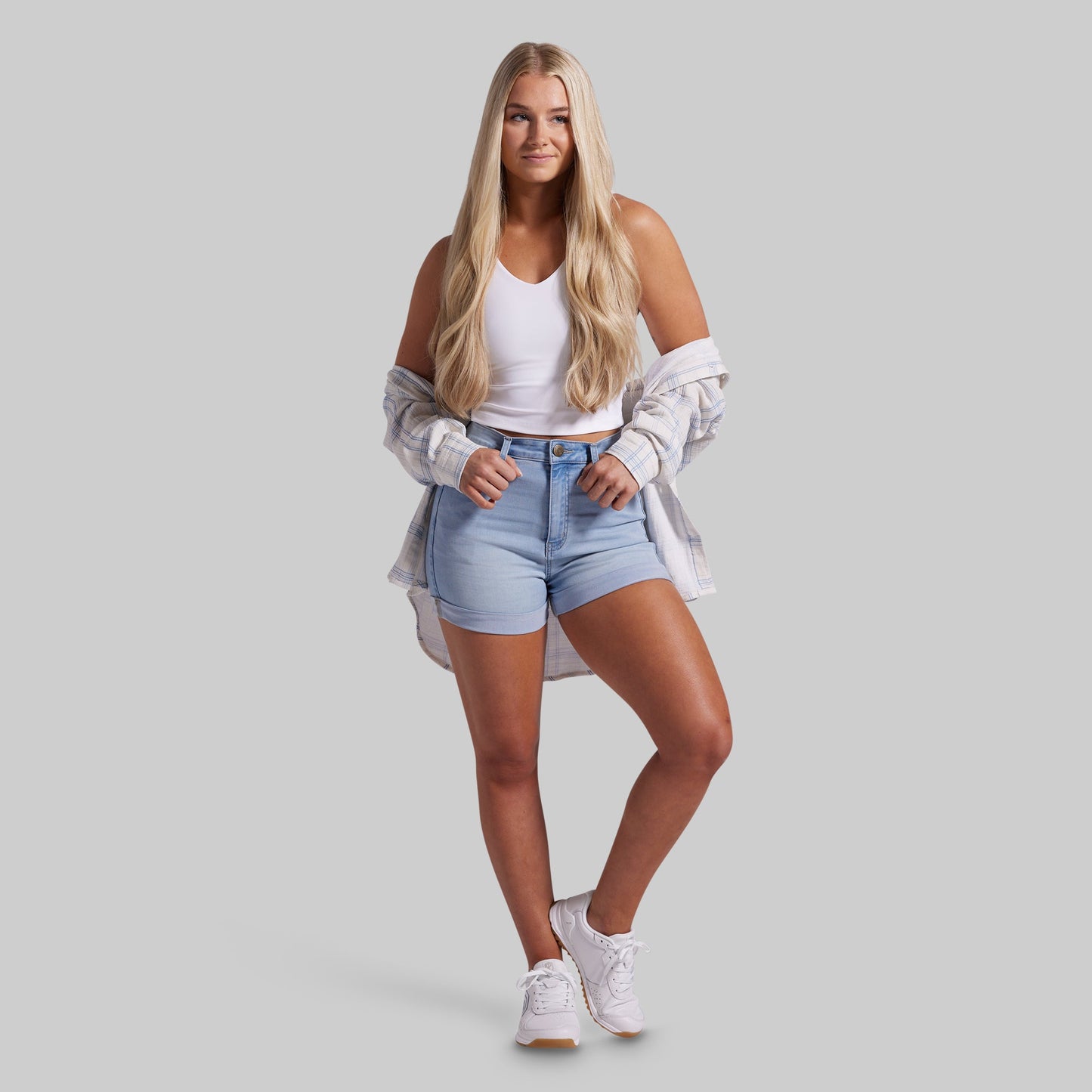 Woman wearing a white crop top, denim shorts, and white sneakers on a gray background