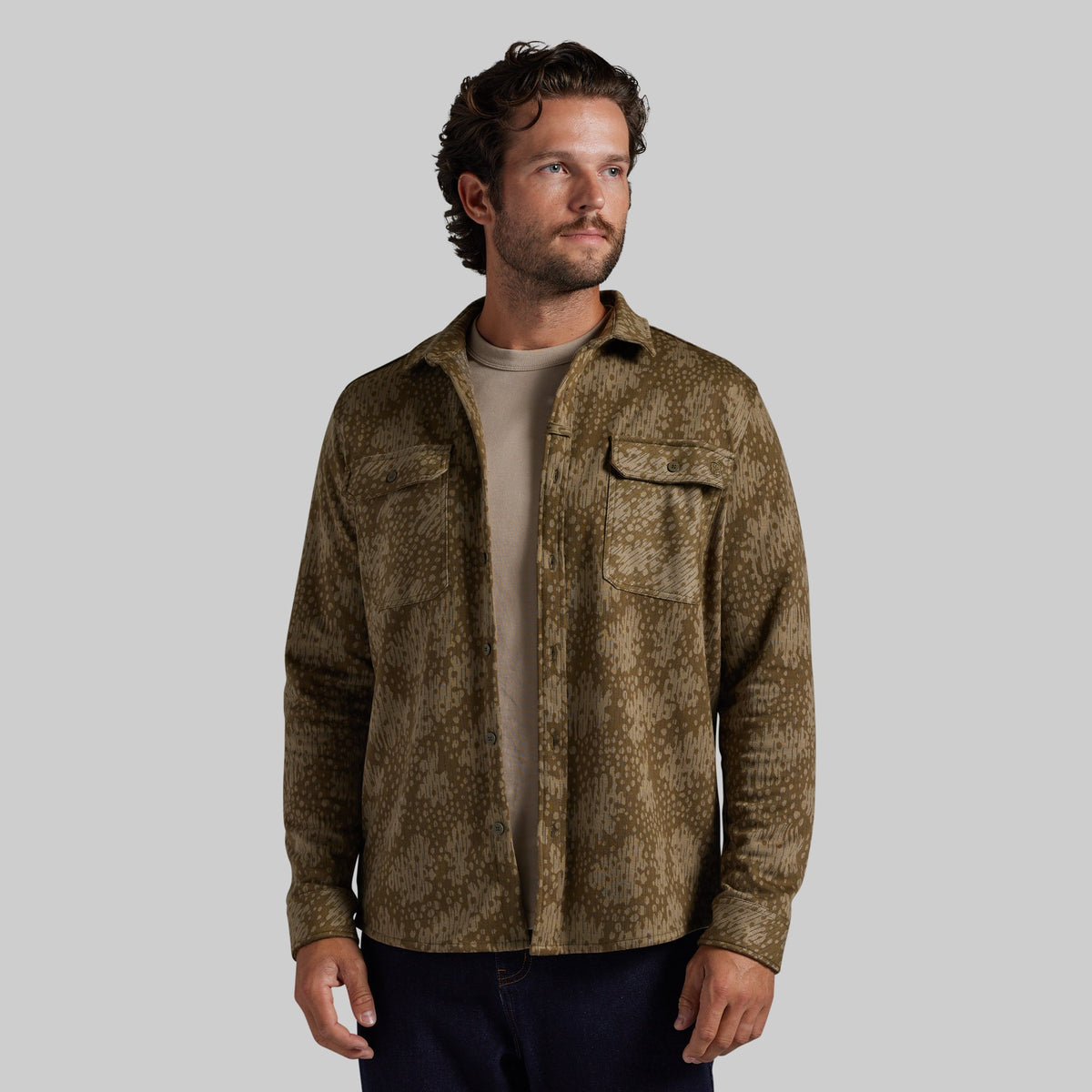 A man wearing a brown and olive green textured camo-print button-up shirt over a beige t-shirt and dark pants, photographed against a solid gray background.