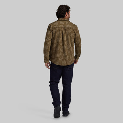 A man viewed from behind wearing a brown and olive patterned button-down shirt, dark navy pants, and black shoes, standing against a solid gray studio background.