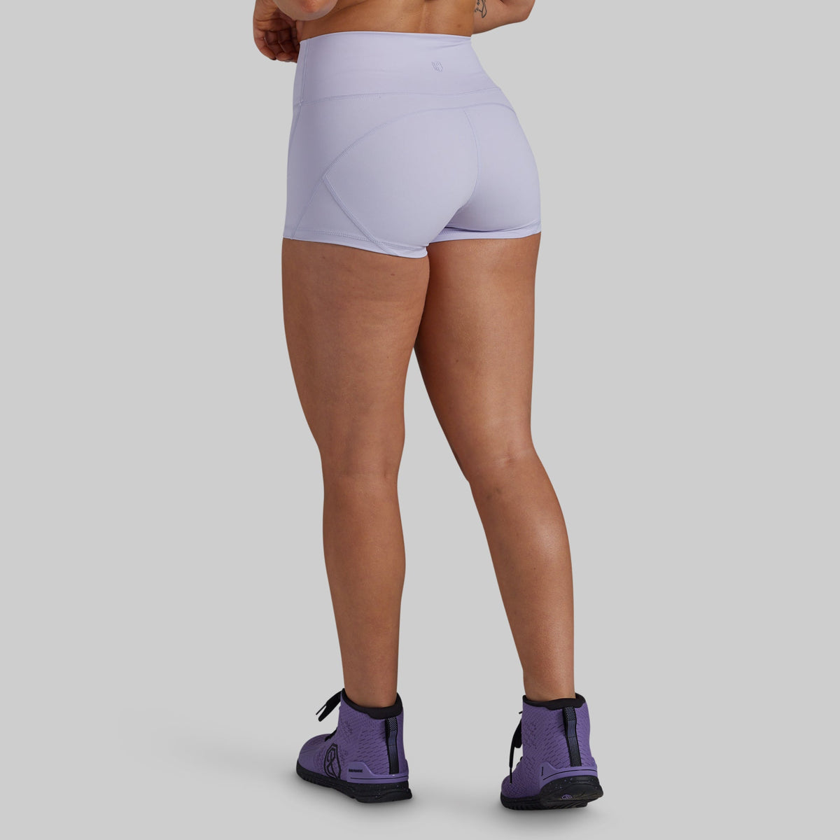 Rear view of a model wearing light lavender high-waisted athletic shorts and purple kettlebell shoes against a solid gray background.