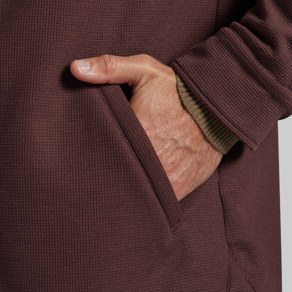 Close-up of a person's hand in a pocket of a textured brown waffle-knit jacket, with a ribbed olive green undershirt cuff visible.
