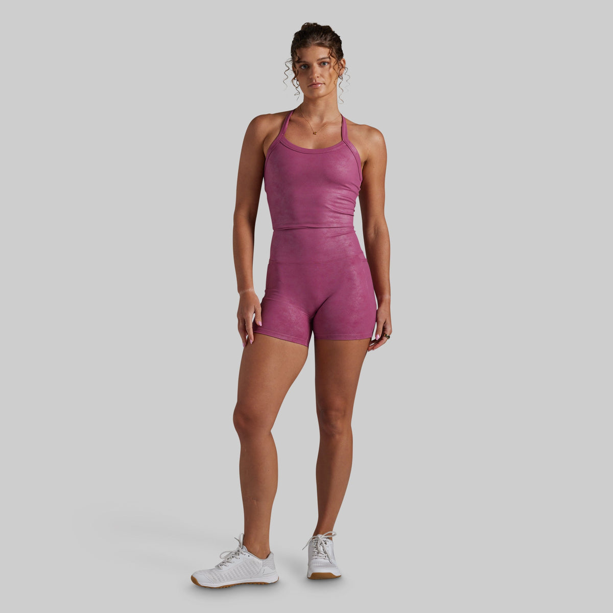 A woman modeling a magenta-pink color athletic set, paired with white sneakers, against a solid light gray background.