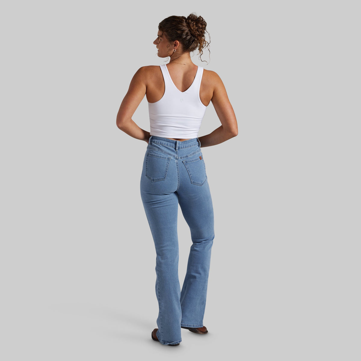 A woman is shown from the back wearing a white tank top and light blue high-waisted bootcut jeans, standing against a solid gray studio background.