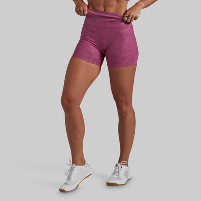 A model wearing high-waisted athletic shorts in a dusty rose or mauve color, paired with white sneakers, shown from the waist down against a solid gray background.
