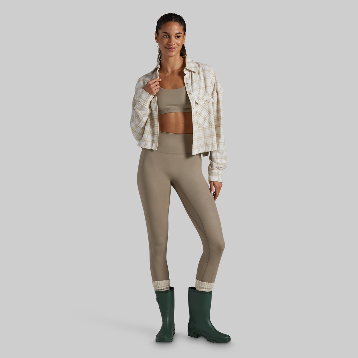 A model stands against a light gray studio background wearing a beige and white plaid button-up shirt over a matching beige sports bra and high-waisted leggings, with dark green rain boots.