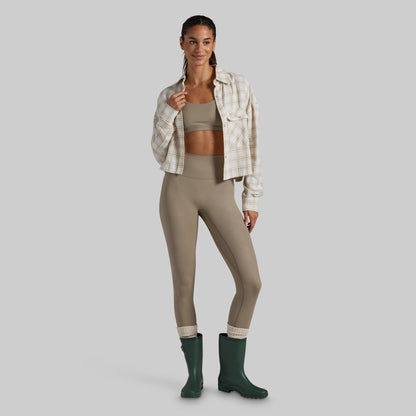 A model stands against a light gray studio background wearing a beige and white plaid button-up shirt over a matching beige sports bra and high-waisted leggings, with dark green rain boots.
