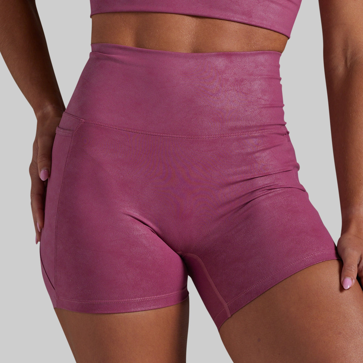 A close-up of a model wearing high-waisted athletic shorts in a dusty rose or mauve color, paired with a matching extended sports bra.