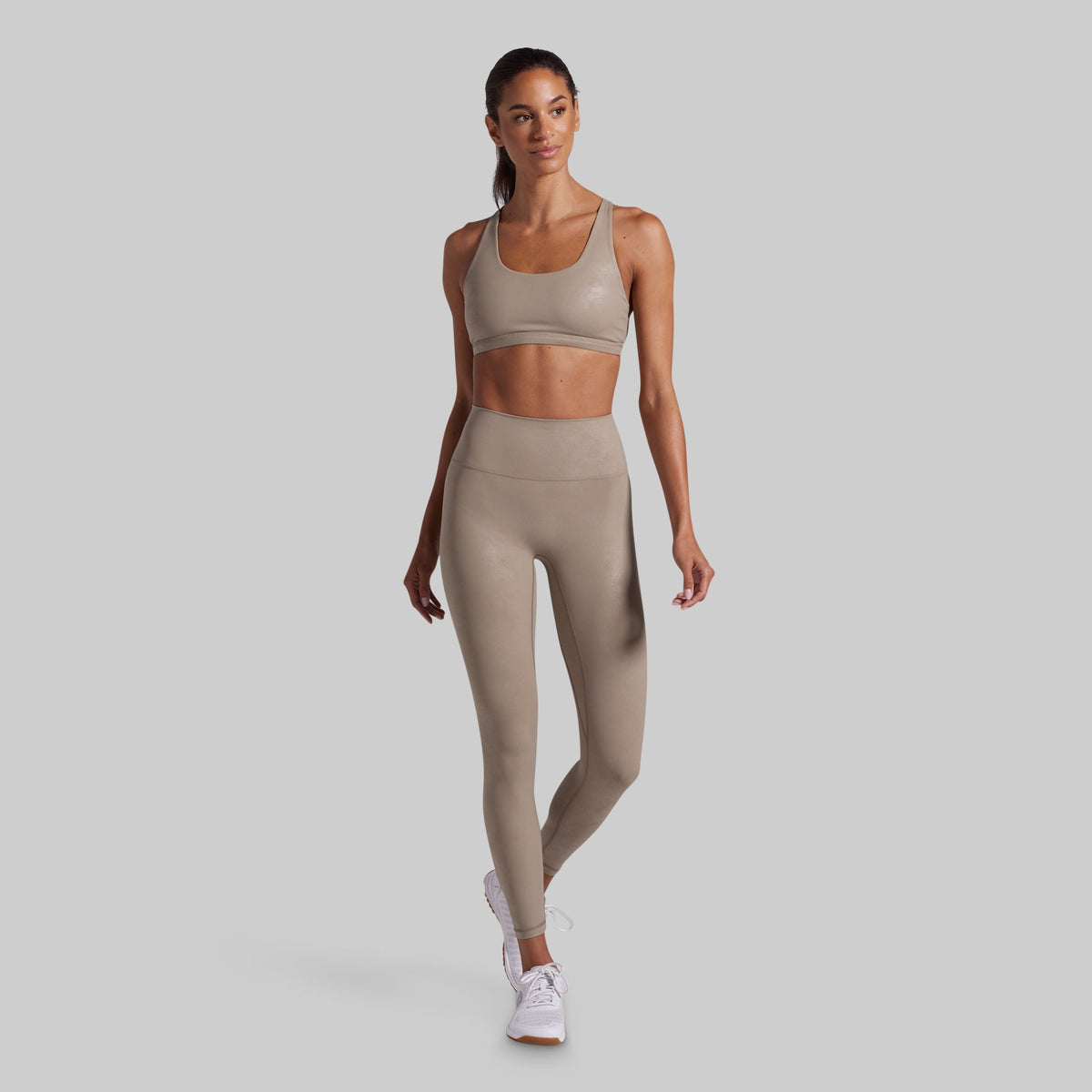 A woman modeling a matching beige activewear set consisting of a sports bra and high-waisted leggings, paired with white sneakers, against a solid light gray background.