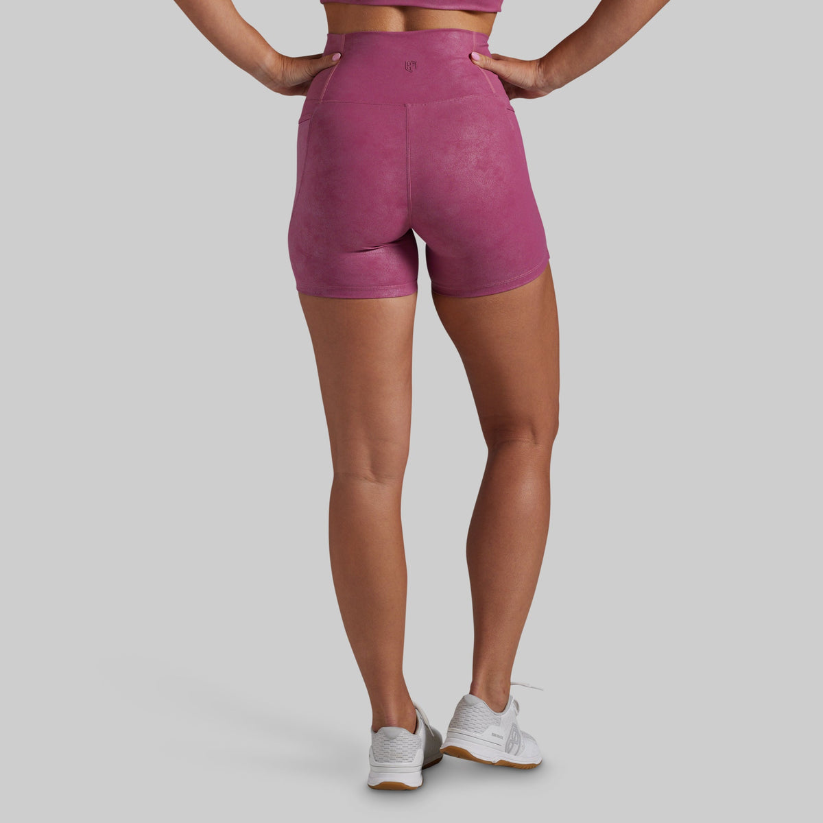 Rear view of a model wearing high-waisted athletic shorts in a dusty rose pink color, paired with white sneakers, against a solid gray background.