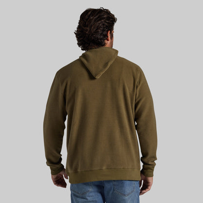 A man viewed from behind wearing an olive green pullover hoodie and blue jeans, standing against a solid light gray background.