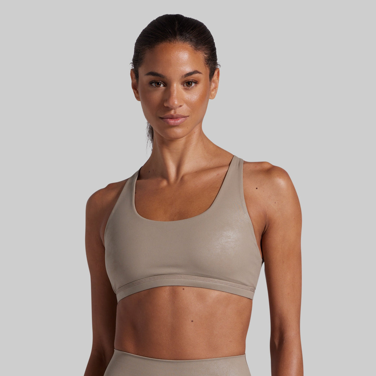 A woman modeling a scoop-neck athletic sports bra in a light taupe-beige color, shown against a solid light gray background.