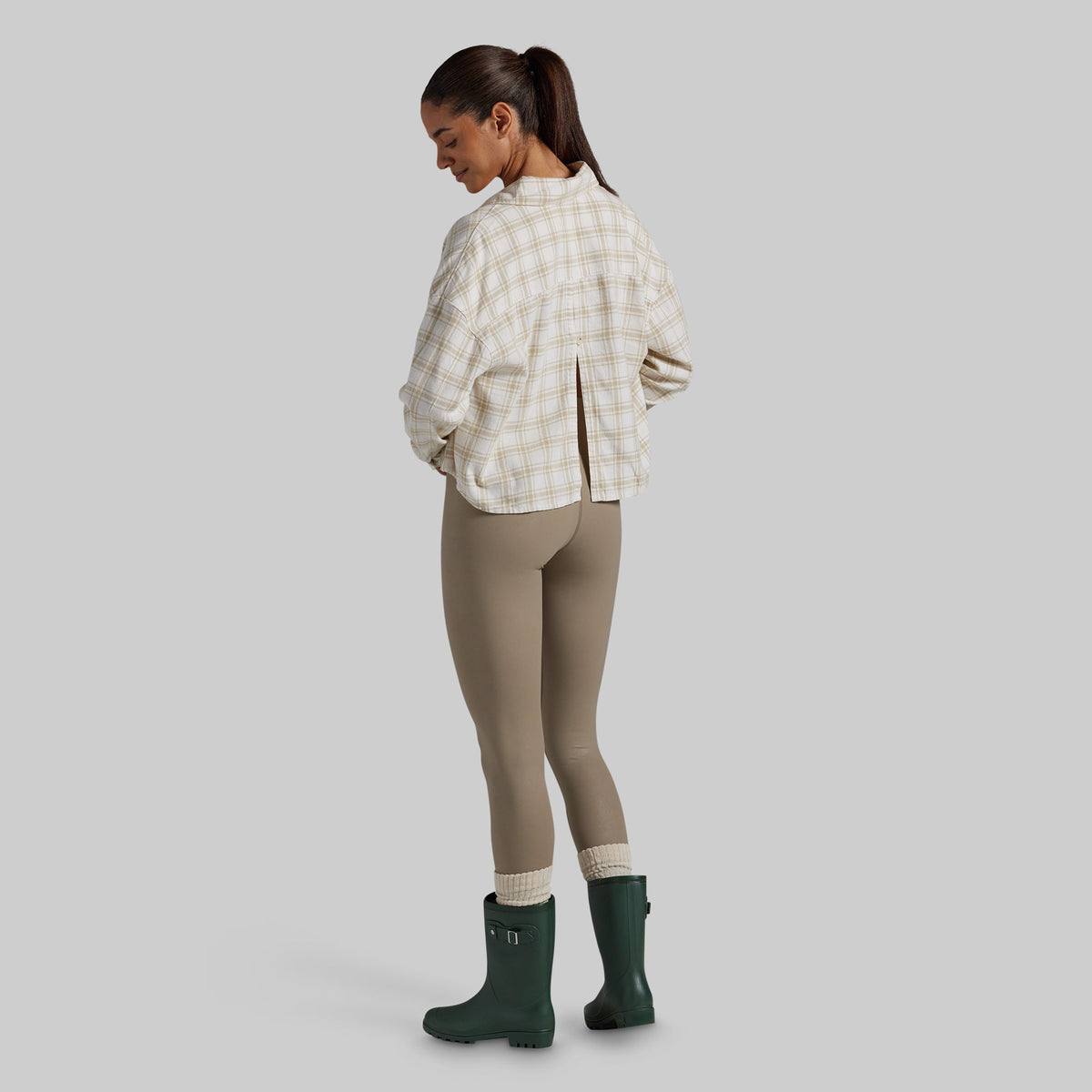 A woman shown from the back wearing a cream and tan plaid cropped overshirt, leggings, and dark green rain boots against a solid gray studio background.