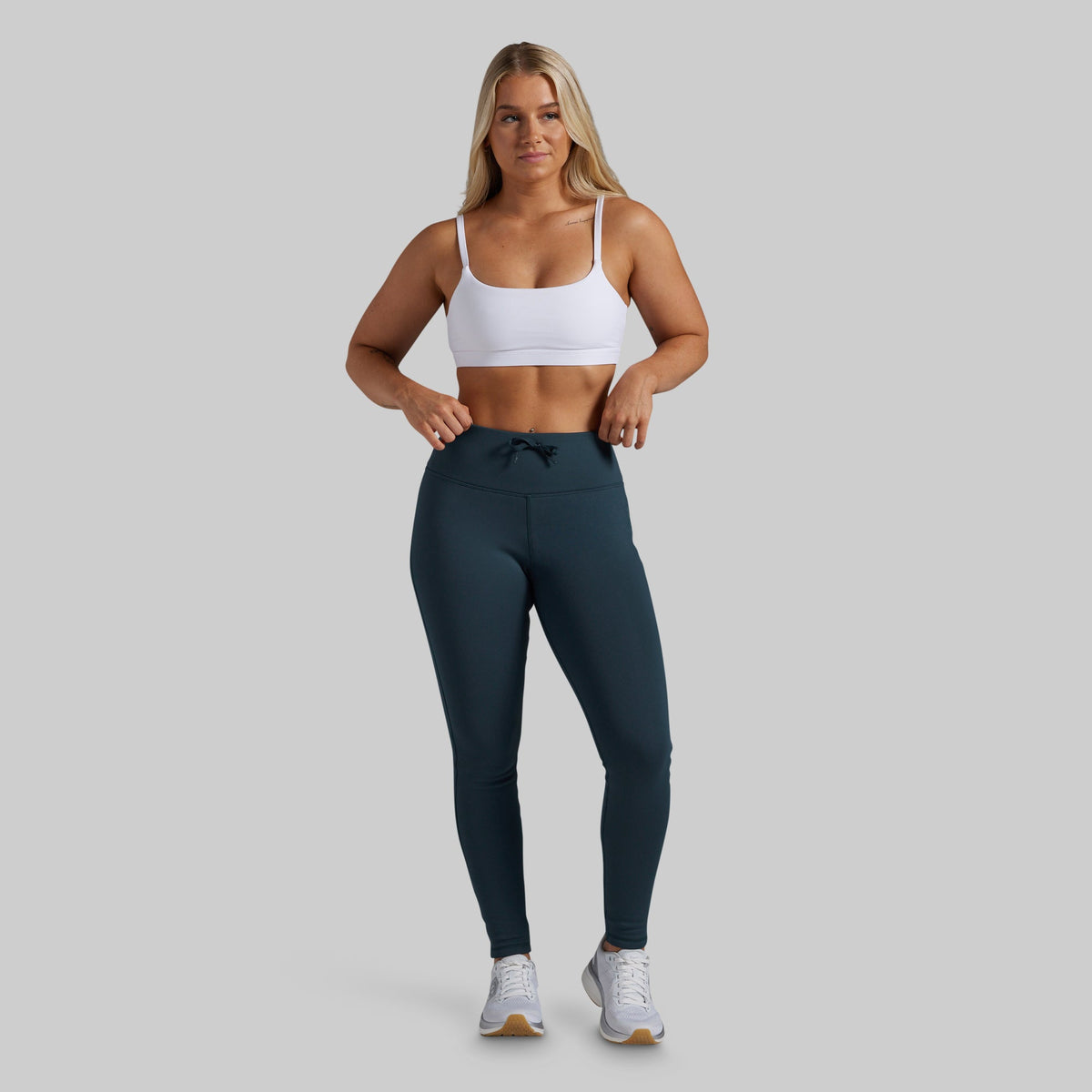 A woman modeling high-waisted athletic leggings in a dark teal color, paired with a white sports bra and white sneakers, against a solid light gray background.