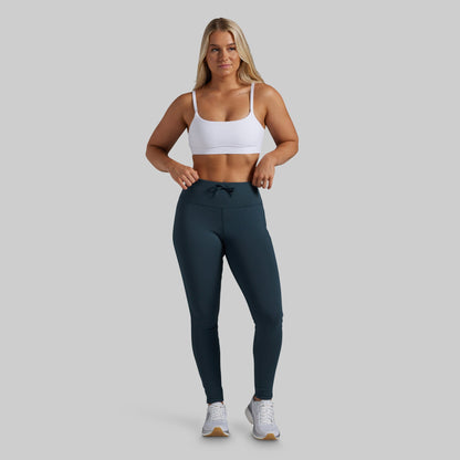 A woman modeling high-waisted athletic leggings in a dark teal color, paired with a white sports bra and white sneakers, against a solid light gray background.
