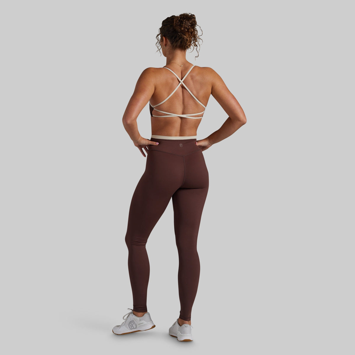 A woman shown from the back wearing a brown athletic leggings set with a matching sports bra featuring cream-colored crisscross straps, posed against a solid light gray background.