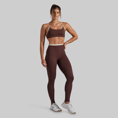 A woman modeling a matching athletic set consisting of a dark brown sports bra and high-waisted leggings with a contrasting beige waistband, paired with white sneakers, against a solid gray background.