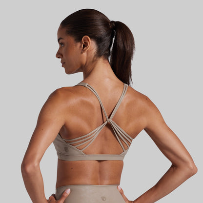 A woman shown from the back wearing a taupe-beige athletic sports bra with multiple thin crossover straps and matching high-waisted leggings, against a solid light gray studio background.