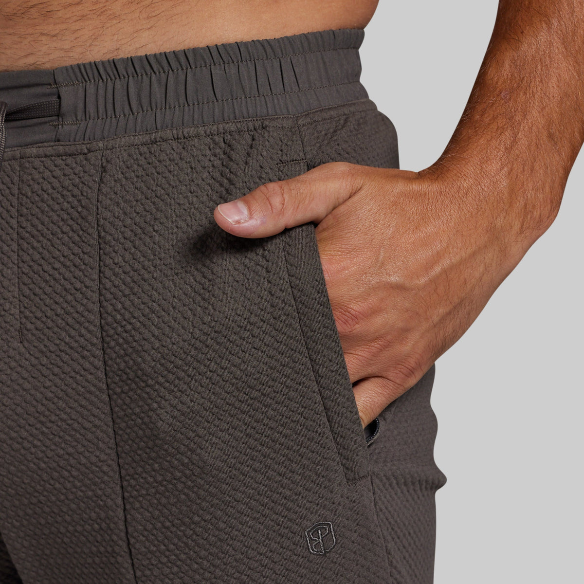 Close-up of a person wearing textured dark gray athletic joggers, showing the side pocket with a hand inserted and an elastic waistband.