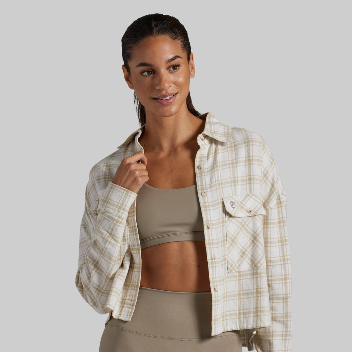 A woman modeling a cropped, long-sleeve plaid button-up shirt in beige and white, worn open over a taupe sports bra and matching leggings, against a solid gray background.