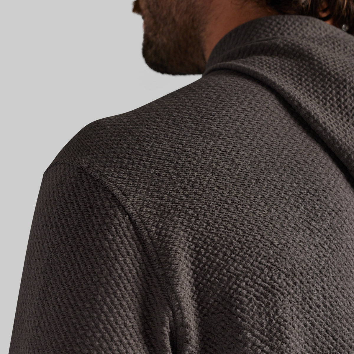 Close-up of a man wearing a dark gray waffle-knit hooded sweatshirt, showing the textured fabric and collar from a side-back angle against a light gray background.