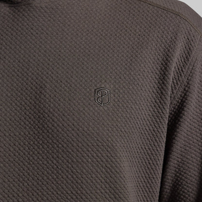 Close-up of a dark grey textured hoodie with a small tonal embroidered shield logo on the chest.