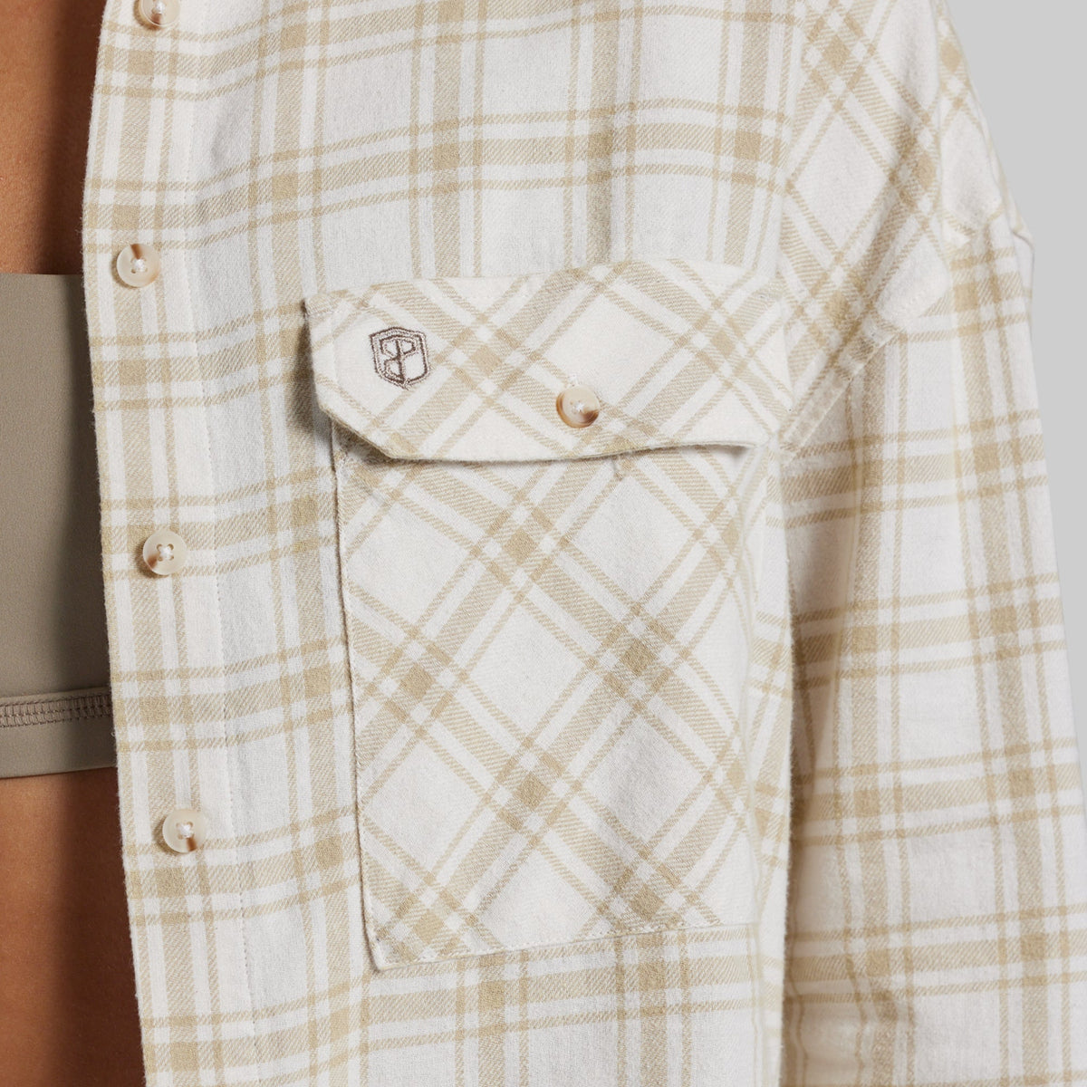 Close-up of a long-sleeve plaid button-up shirt in cream and tan, featuring a chest pocket with a buttoned flap and a small shield logo patch.