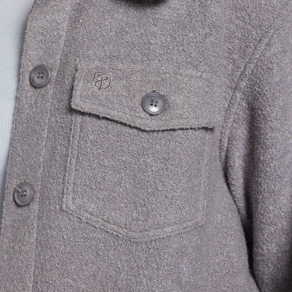 Close-up of a textured gray button-up jacket with a chest pocket, showing a small shield logo on the pocket flap.