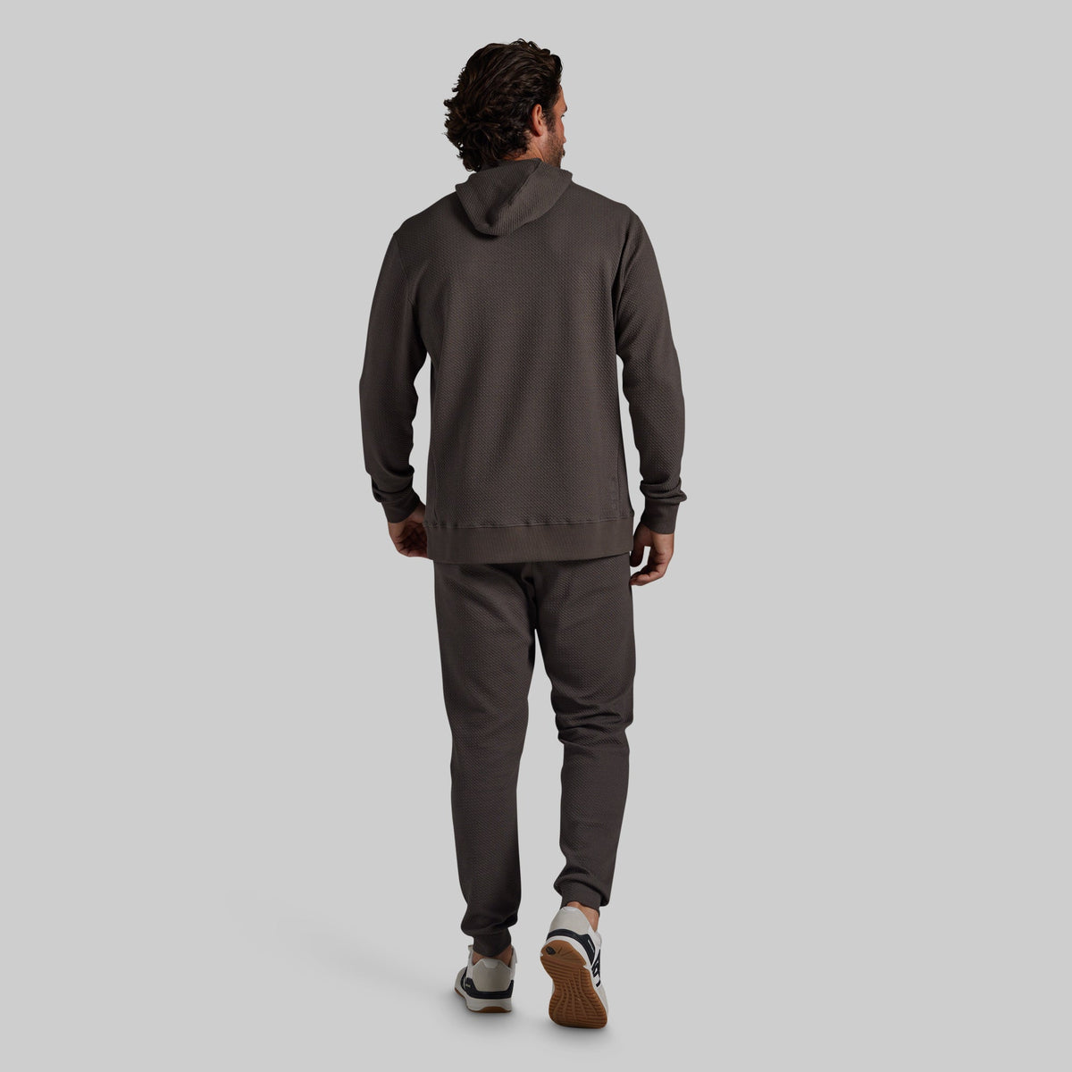 A man is shown from behind wearing a dark grey hooded sweatshirt and matching jogger pants, walking against a solid light gray background.
