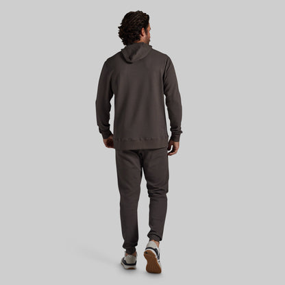 A man is shown from behind wearing a dark grey hooded sweatshirt and matching jogger pants, walking against a solid light gray background.
