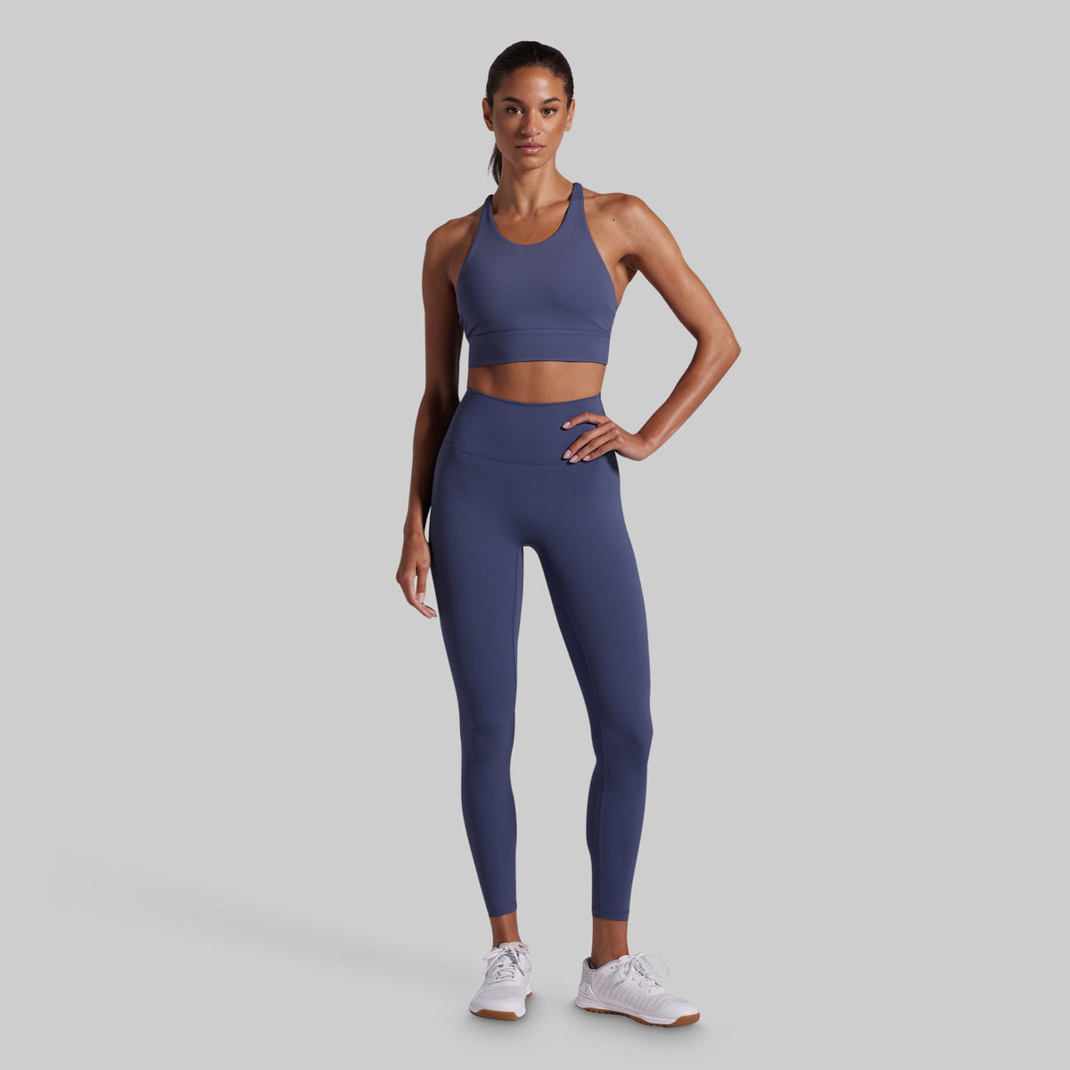 A woman modeling a matching athletic set consisting of a blue sports bra and high-waisted leggings, paired with white sneakers, against a solid gray background.