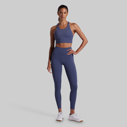 A woman modeling a matching athletic set consisting of a blue sports bra and high-waisted leggings, paired with white sneakers, against a solid gray background.