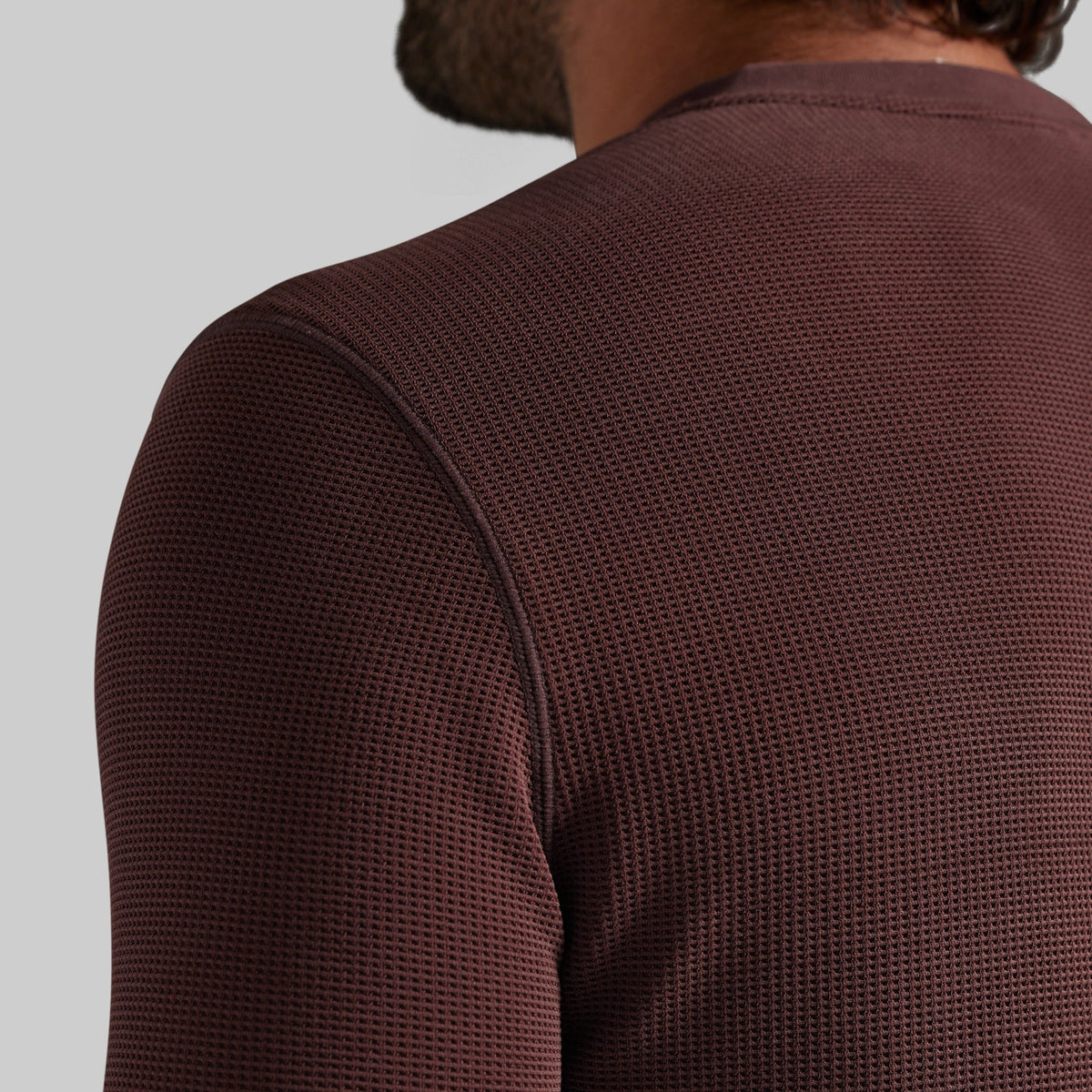 Close-up back view of a person wearing a textured, waffle-knit long-sleeve shirt in a deep burgundy brown color, set against a solid light gray background.