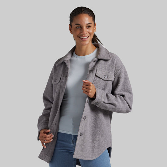 A woman wearing a heather gray button-up jacket over a light blue top and blue jeans, standing against a solid gray studio background.