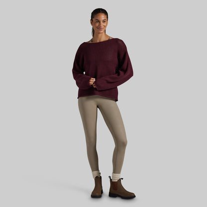 A woman modeling a dark burgundy ribbed knit sweater with a wide boat neckline, paired with beige leggings and brown Chelsea boots, against a solid light gray background.