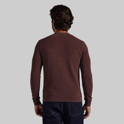 A man viewed from behind wearing a dark brown long-sleeve crewneck shirt and dark blue jeans, set against a solid light gray background.