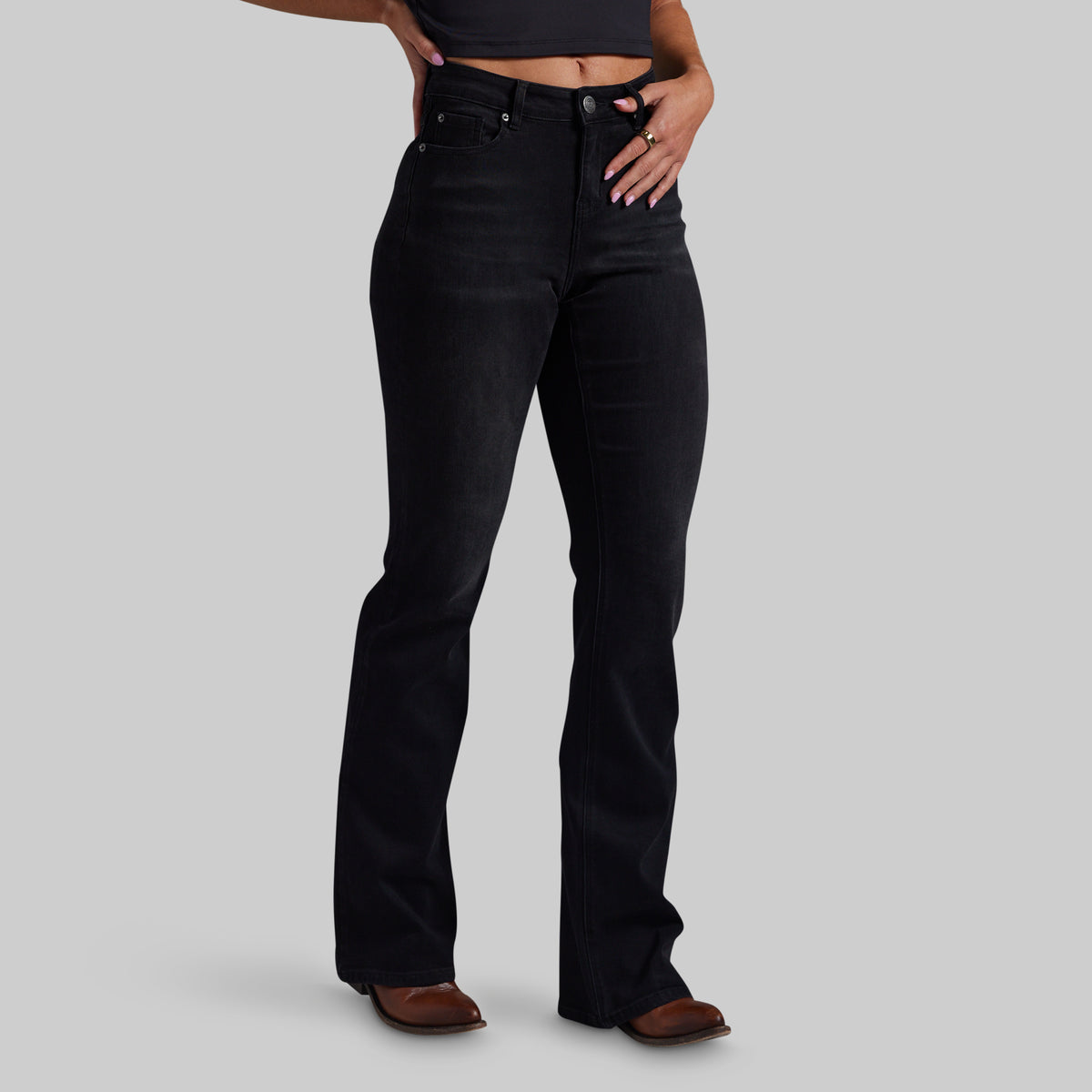A model wearing black mid-waisted bootcut jeans and brown leather shoes, shown from the waist down against a solid gray studio background.