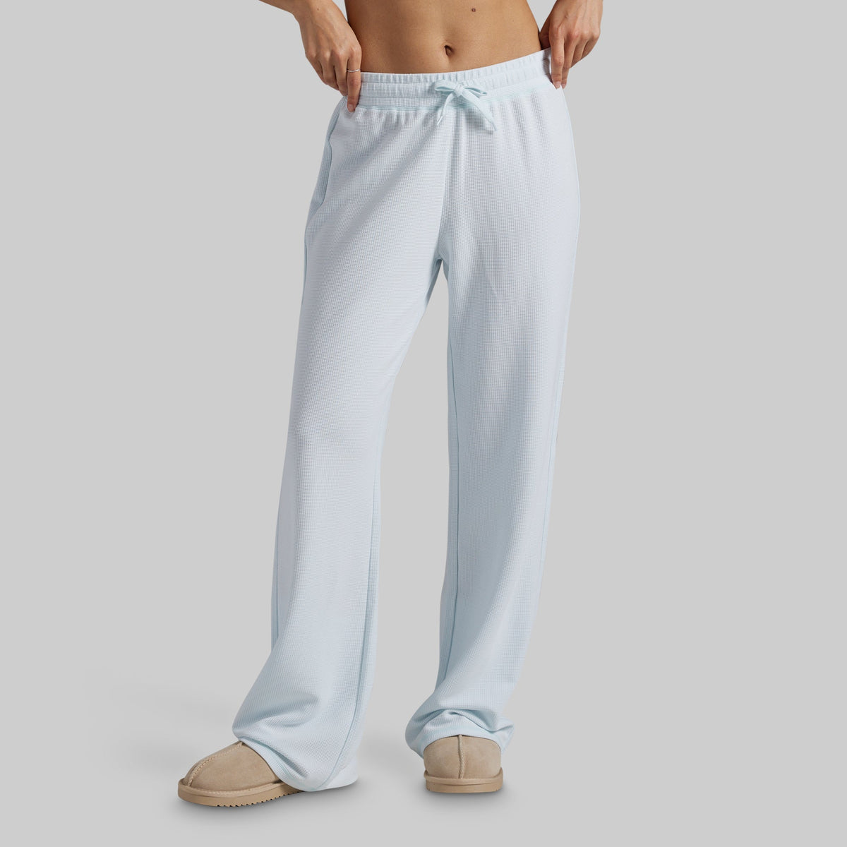 A person wearing light blue wide-leg lounge pants with a drawstring waistband and beige slippers, shown from the waist down against a plain light gray studio background.